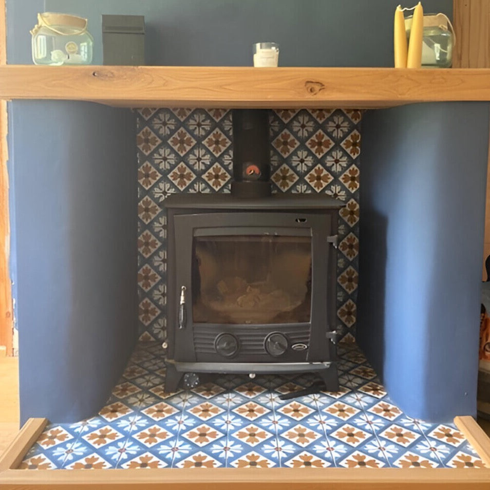 A cozy fireplace setup featuring a wood-burning stove in a dark blue room. The stove is set on a patterned tile hearth with blue and white tiles featuring a star and floral design. Above the stove, a wooden mantel holds decorative items including candles and a jar. The stovepipe extends up into the wall, partially hidden by the mantel.